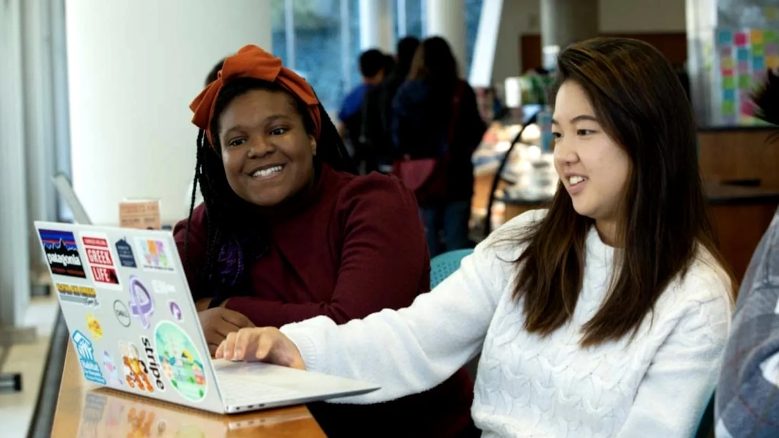 Two CMU students sitting at a table looking at a laptop covered in stickers, smiling while in conversation about the FAFSA