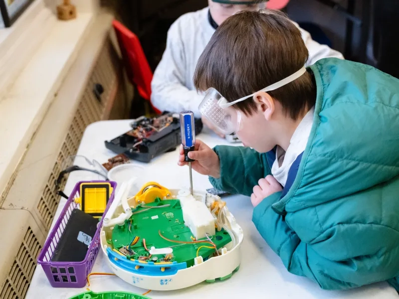 Child wearing a teal puffer coat and safety goggles plays with a screwdriver and circuit board.