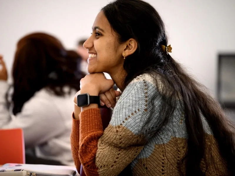A CMU student wearing a colorful sweater and a smartwatch is smiling while seated in a classroom, attentively listening