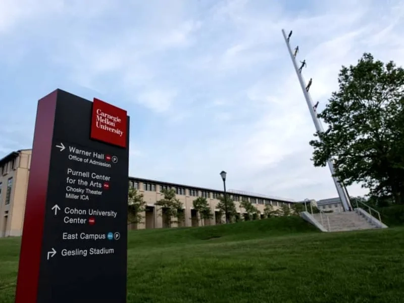 Carnegie Mellon University directional signpost for campus visits on green grass with buildings and a sculpture in the background