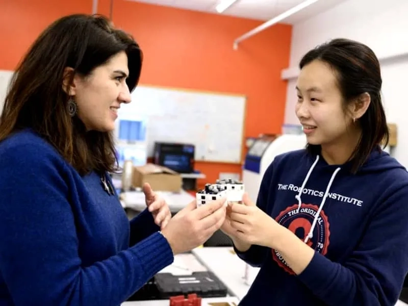 A CMU student and professor talk as the student holds a small tech gadget while both are standing in a classroom workshop