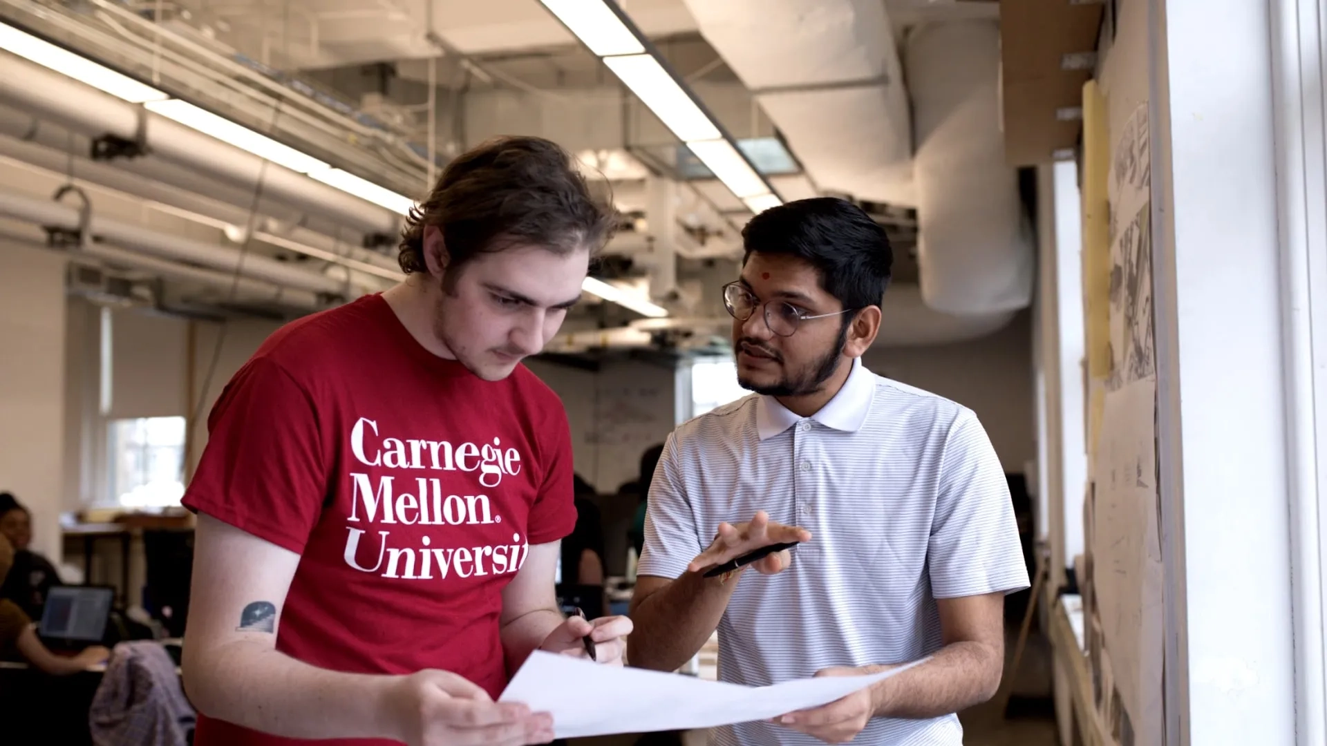 A student wearing a red Carnegie Mellon University t-shirt holds papers while talking to another student in a CMU classroom