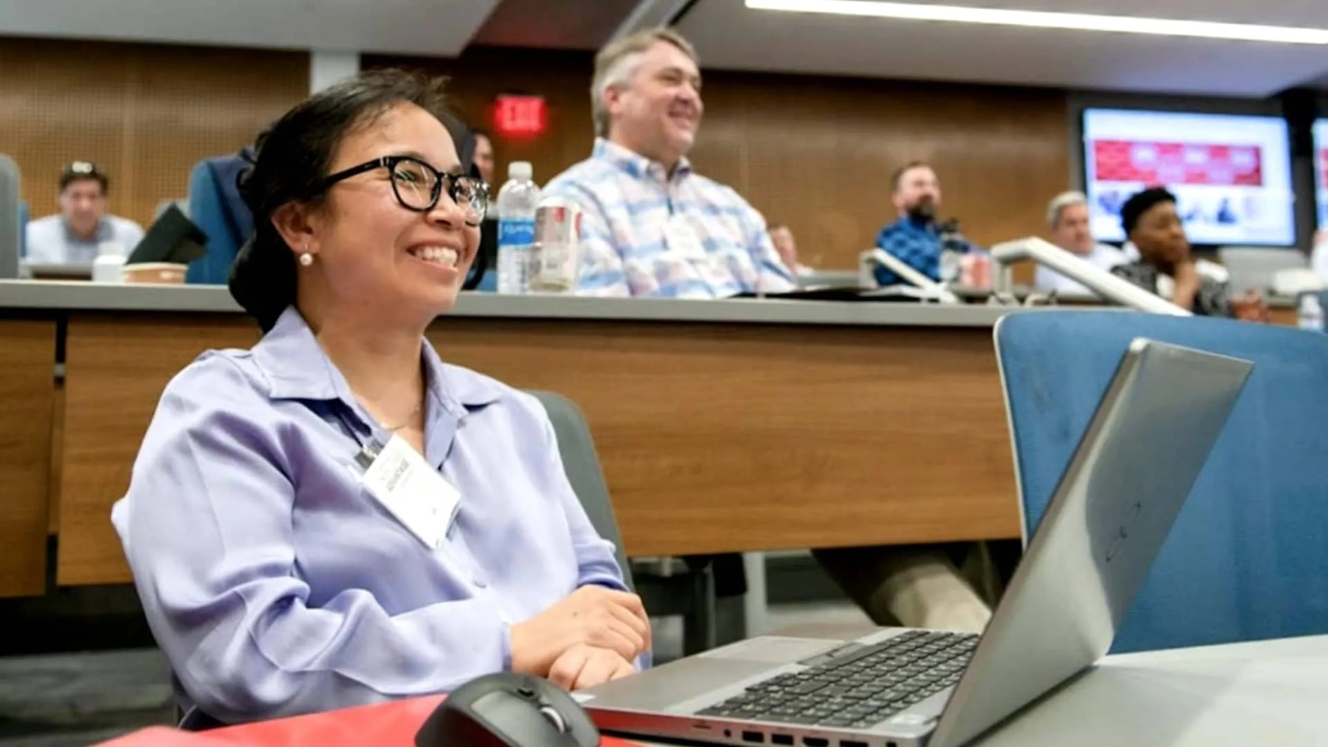 A CMU continuing education student smiling while seated in a classroom with a laptop on a desk and other students in the background