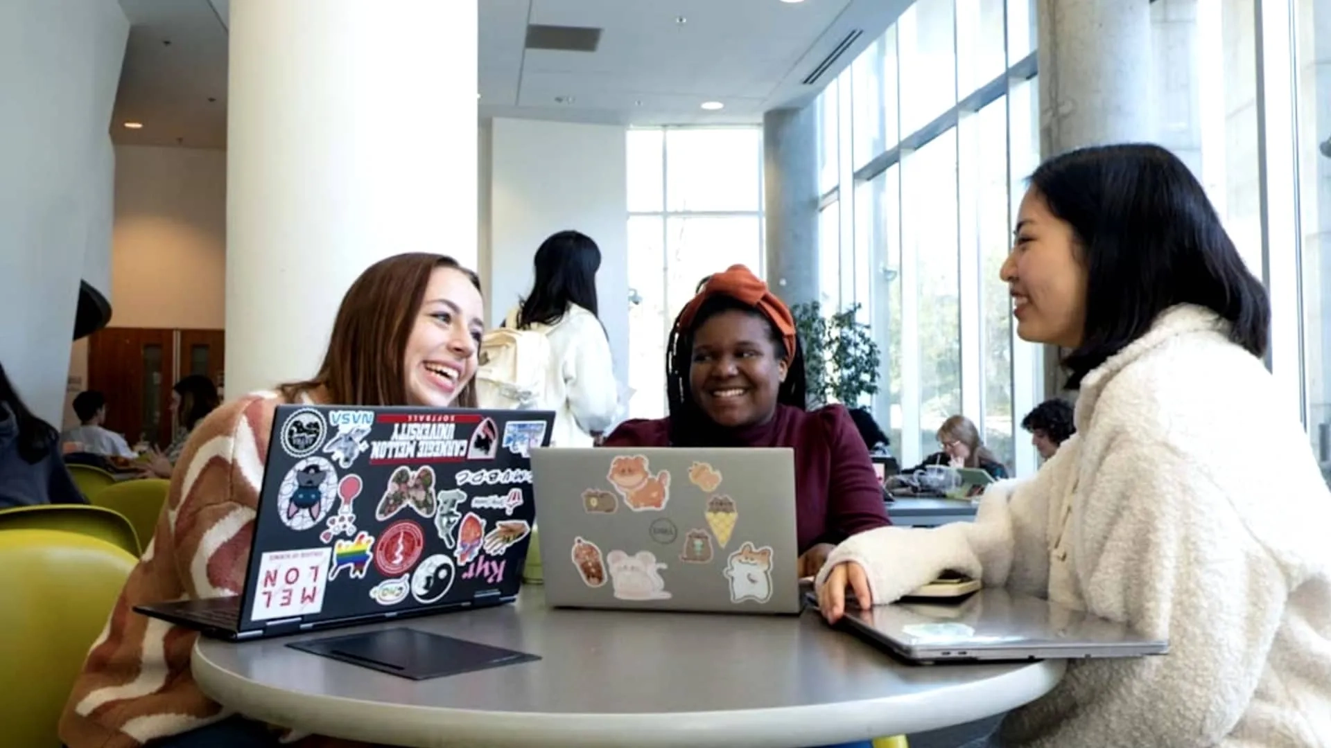 Three CMU students sit at a round table in the student center, smiling, talking, and working on their sticker-covered laptops