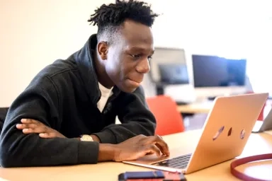 A CMU student wearing a black jacket is focused on working at a laptop in a classroom setting with open computers in the background
