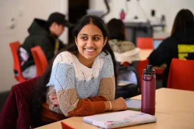A CMU student wearing a colorful sweater smiles while sitting at a desk in a classroom with a laptop and water bottle nearby
