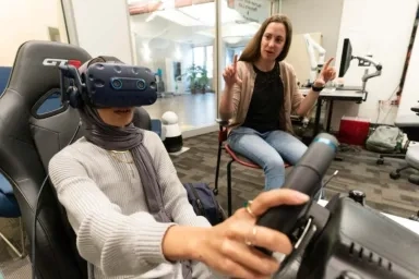 A CMU student uses a virtual reality headset and steering wheel in a simulation, guided by an instructor in a classroom workshop