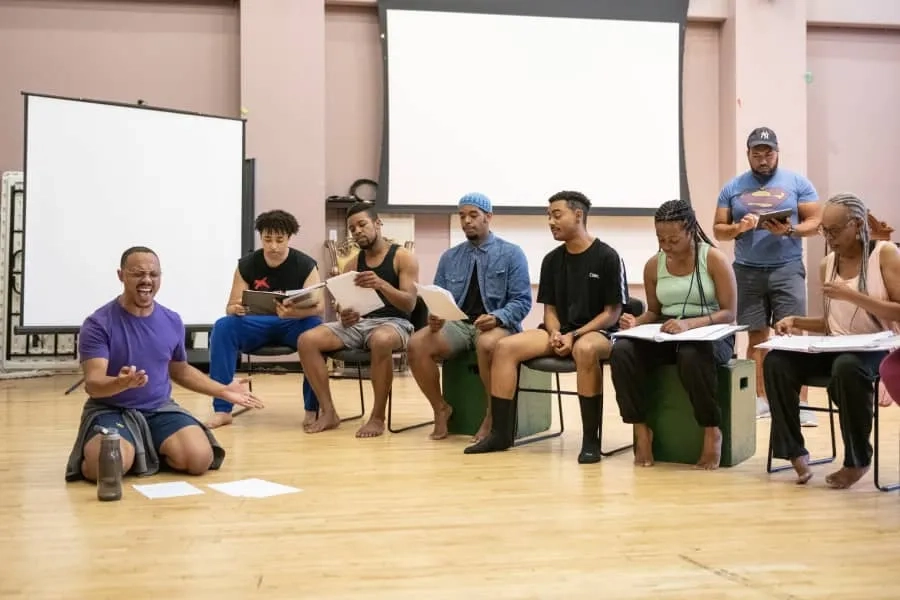 A group of CMU drama students are engaged in rehearsal. One student kneels while others sit  in chairs with scripts in hand.