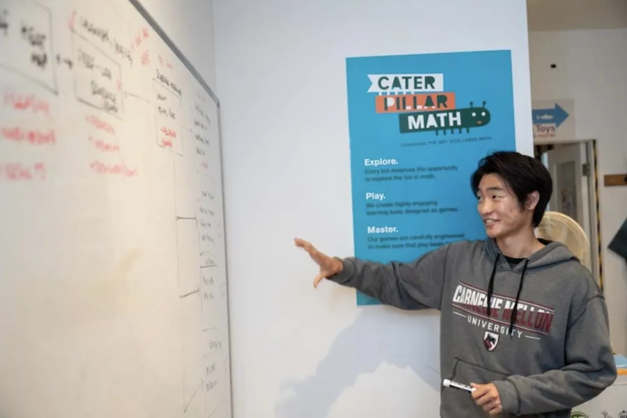 A Carnegie Mellon student wearing a CMU sweatshirt points toward a whiteboard with diagrams and notes