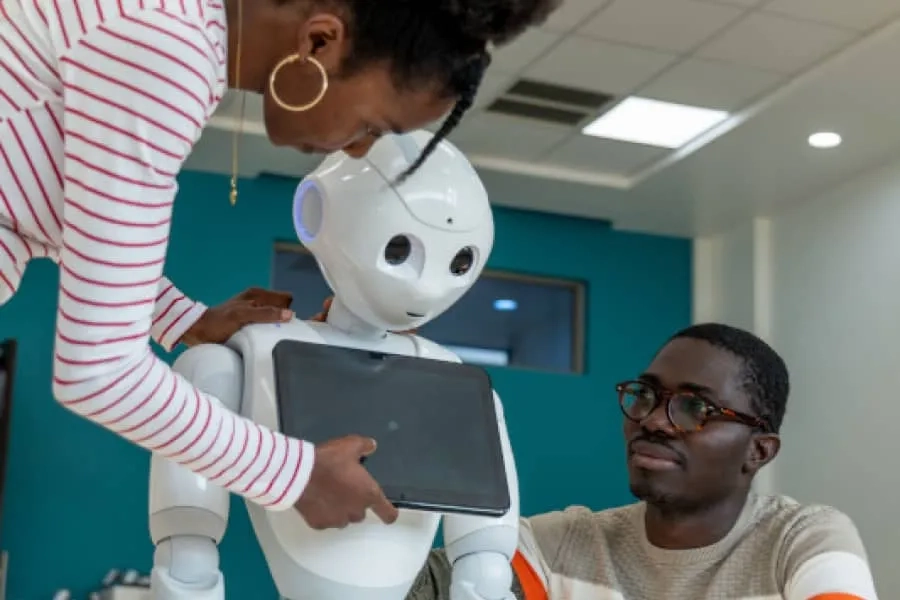 Two CMU students interact with a white human-like robot in a classroom, placing a tablet on the robot's chest.